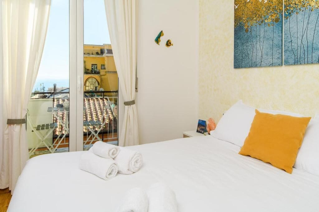 Habitación de hotel con cama, ropa blanca, almohada amarilla y vista a un balcón con ropa tendida.
