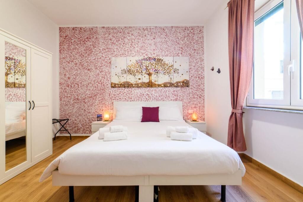 Una cama bien hecha con sábanas blancas y un cojín morado, dos mesitas de noche con velas, un papel tapiz estampado con flores y una ventana con cortinas.