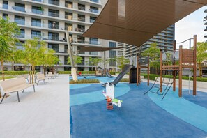 A playground with a slide, swings, and benches in an urban setting with modern buildings in the background. - Stayshort - Elegant 1 Bedroom in Emaar Beachfront That Sleeps 2 (Dubai)