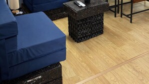 A room with a blue sofa, a wooden dining table, and a black ottoman with a remote control.