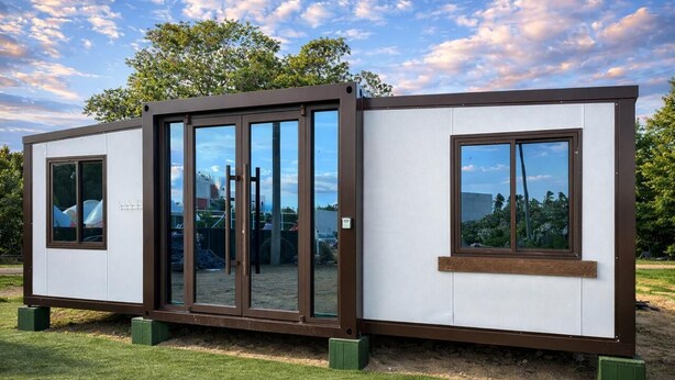 A modern, modular building with large windows and a sliding door, set in a grassy area with trees in the background.