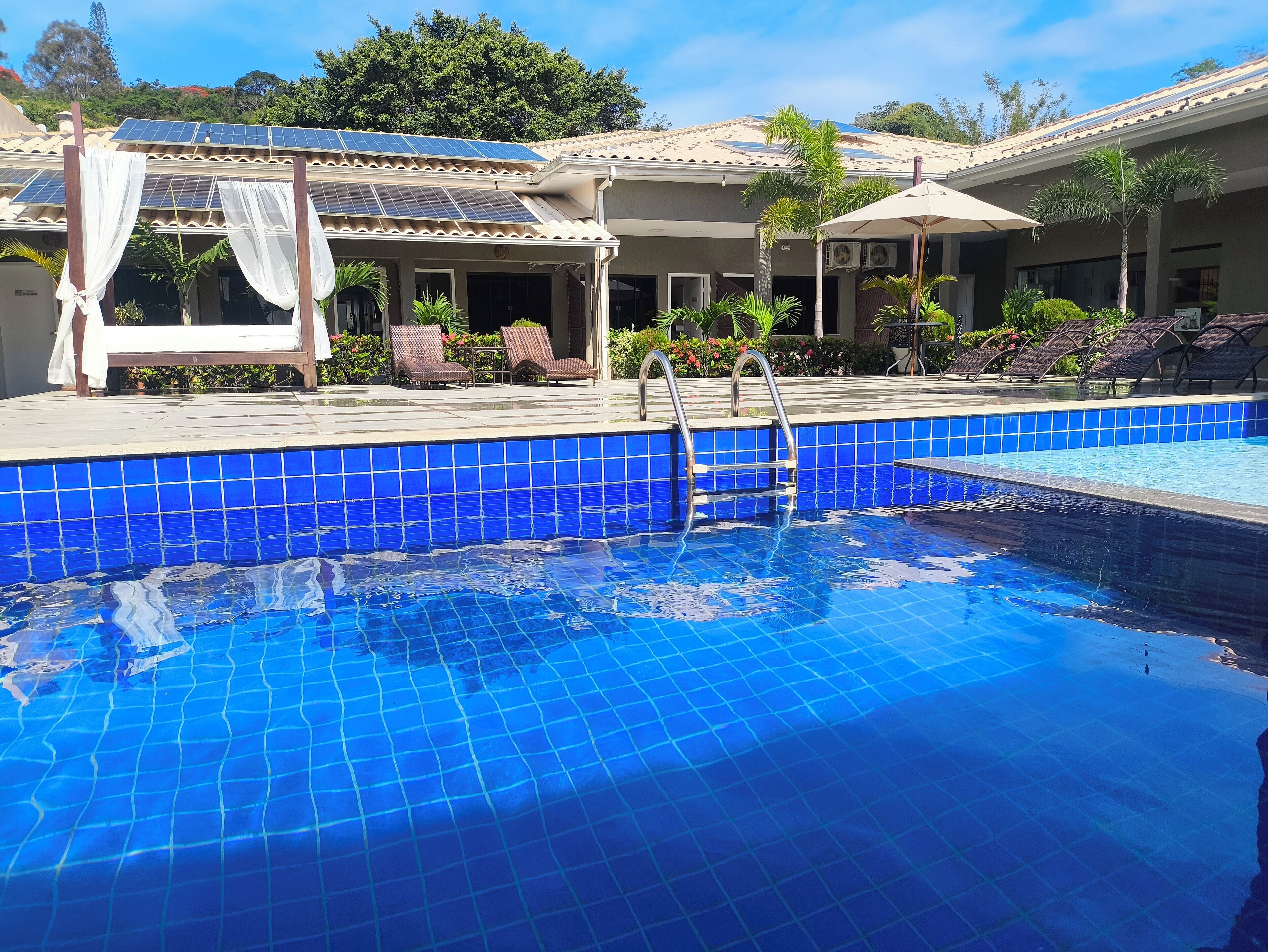Una piscina con baldosas azules, sillas de descanso y una pérgola con cortinas blancas.