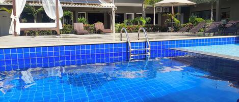 Una piscina con baldosas azules, sillas de descanso y una pérgola con cortinas blancas.