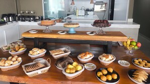 Une table de buffet garnie de différents types de pain, de pâtisseries, et d’un gâteau au premier plan, avec une cuisine visible à l’arrière-plan.