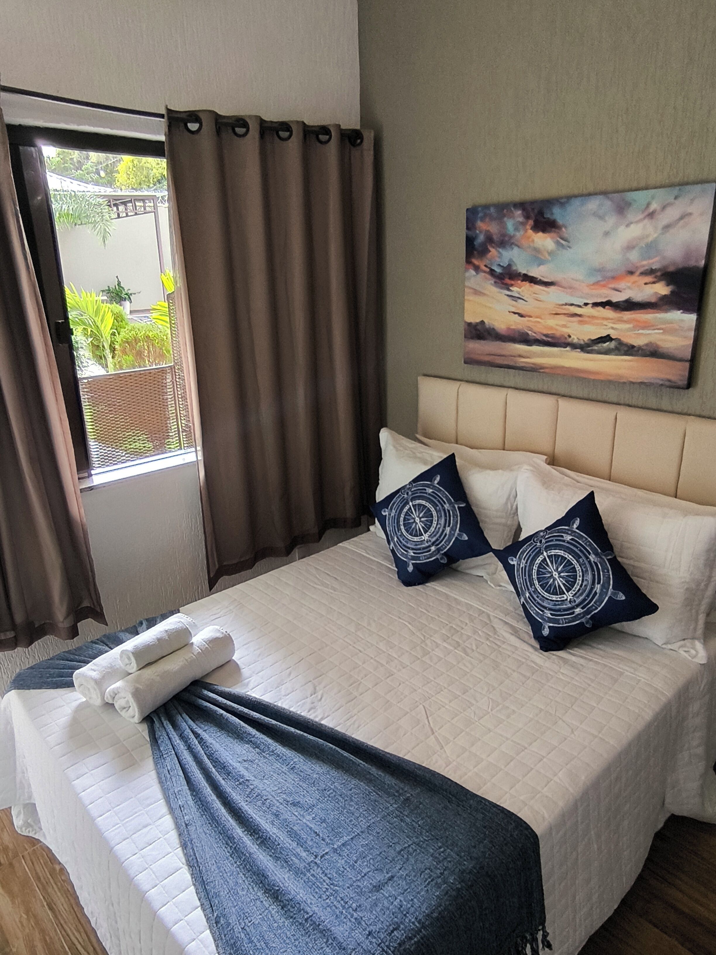 A bedroom with a bed, pillows, a blanket, a painting, and a view of greenery outside.