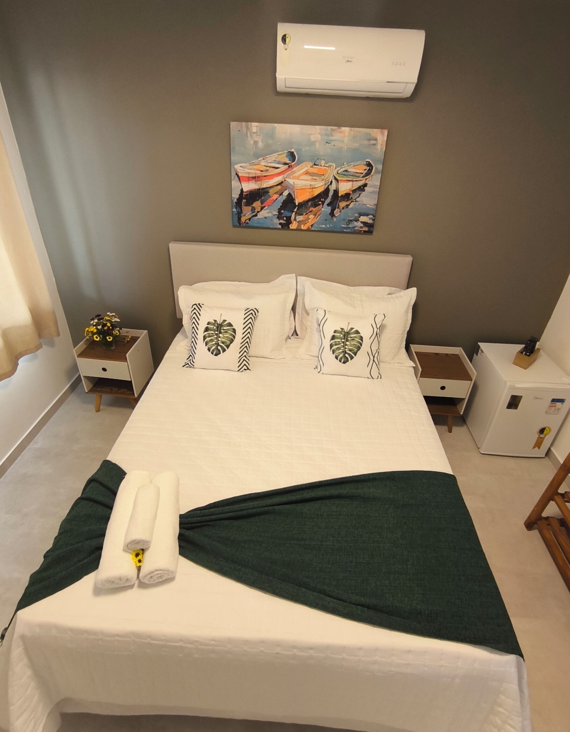 A neatly made bed with a green throw, white pillows, and a painting of boats on the wall.