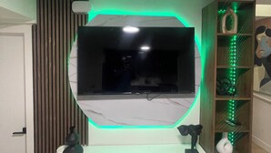 Une chambre moderne équipée d’un téléviseur encastré, d’un bureau blanc avec tiroirs, d’une étagère en bois agrémentée d’objets décoratifs et d’éclairage LED vert.