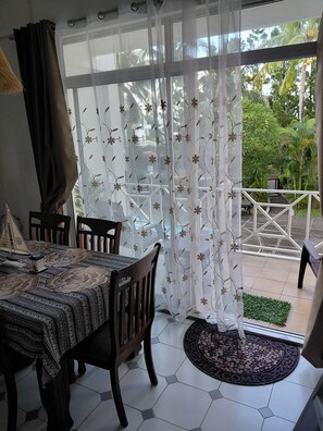 A dining area with a table and chairs, a patterned tablecloth, and a view of the outdoors through sheer curtains.