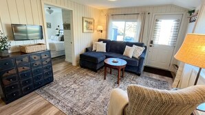 Un salon chaleureux avec un canapé bleu foncé, une table basse ronde et un tapis à motifs. On y trouve un téléviseur posé sur un meuble, une commode avec des tiroirs et une lampe sur une table d’appoint.