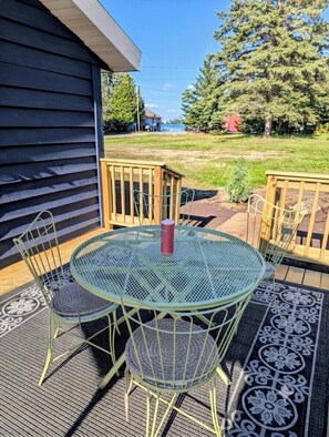 A metal table and chairs on a deck with a patterned rug, a wooden railing, and a view of a grassy area and trees.