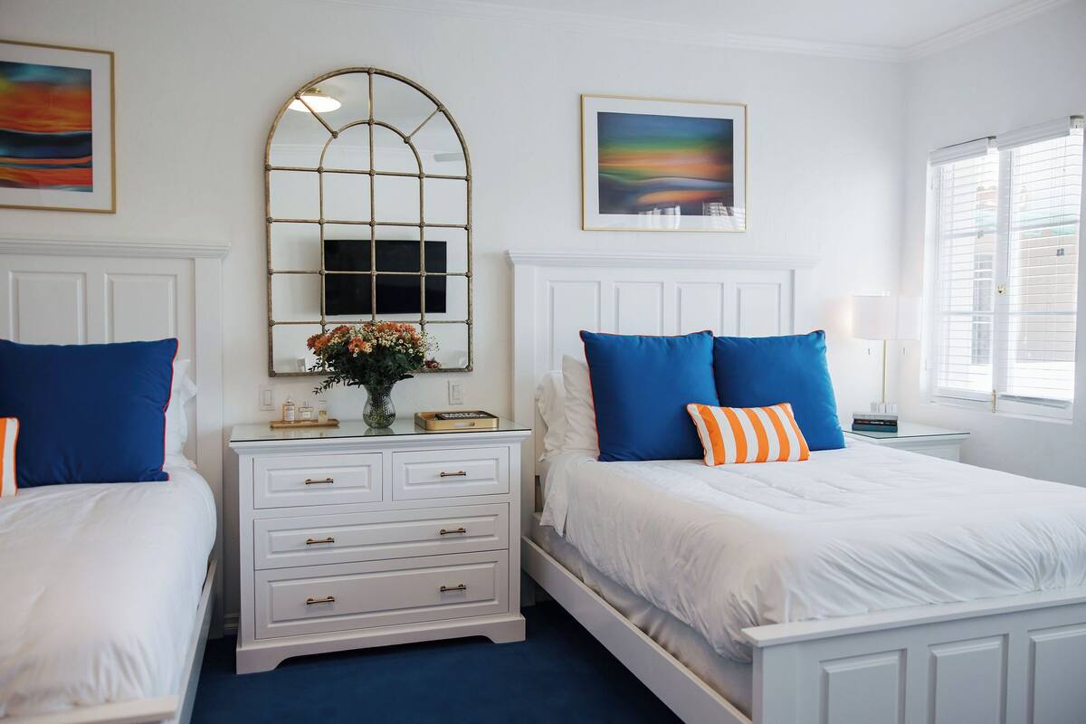 Habitación de hotel con dos camas, cabeceros blancos y almohadas azules. Se observa un espejo grande en la pared y una ventana con persianas.