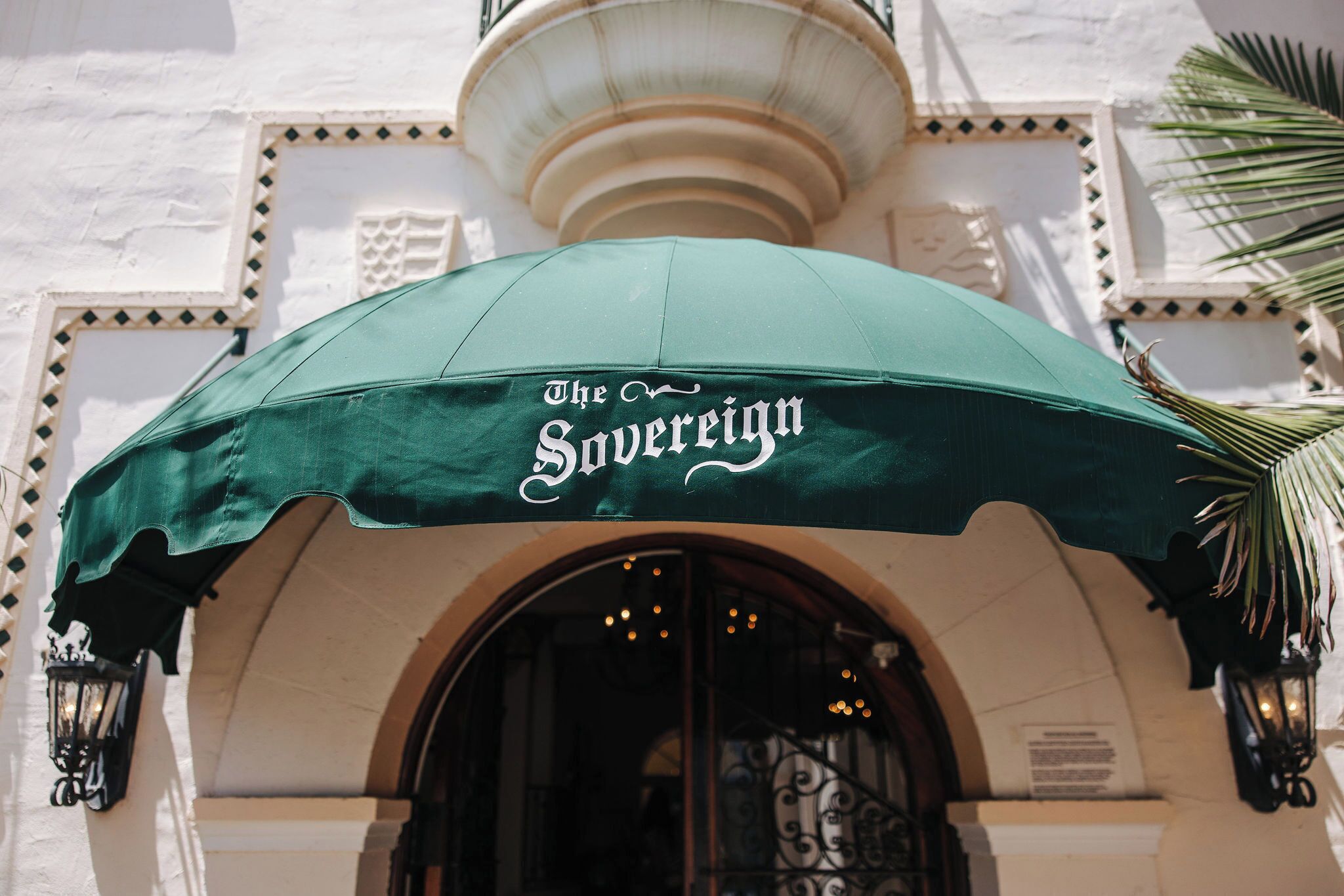 A green awning with "The Sovereign" written on it, an arched entrance, and a palm tree to the right.