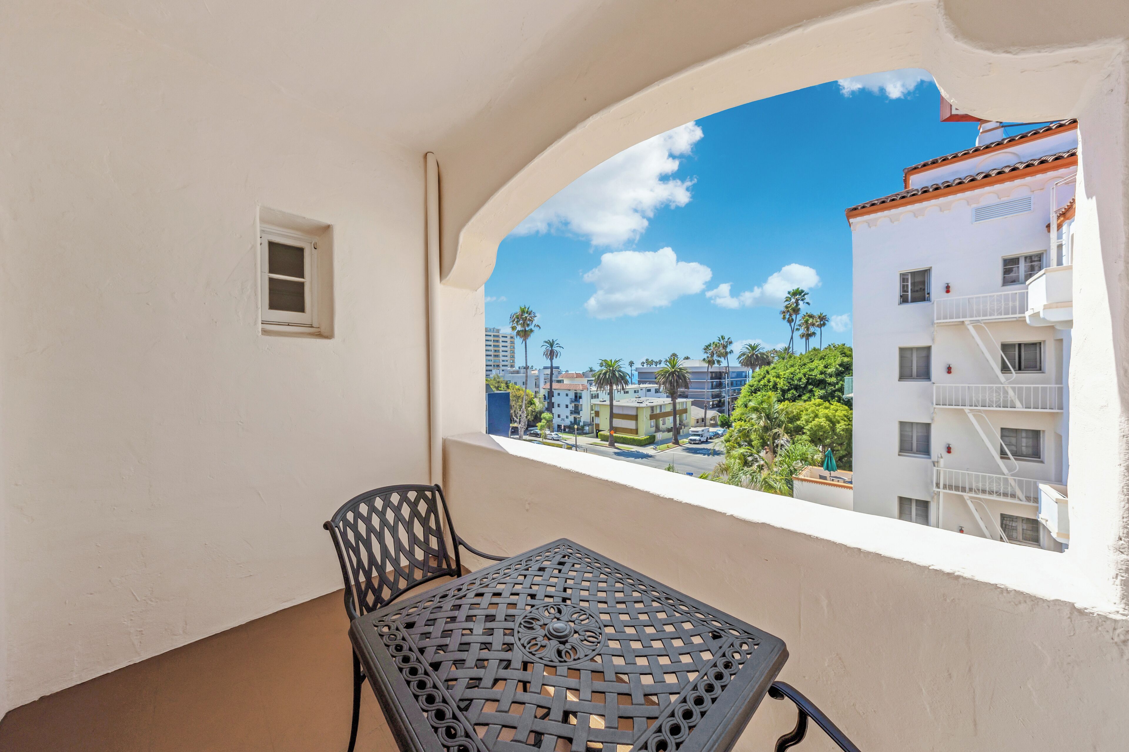A balcony with a table and chair, overlooking a residential area with palm trees and buildings.