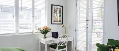 A room with a white desk, a green chair, a framed picture on the wall, and a window with blinds.