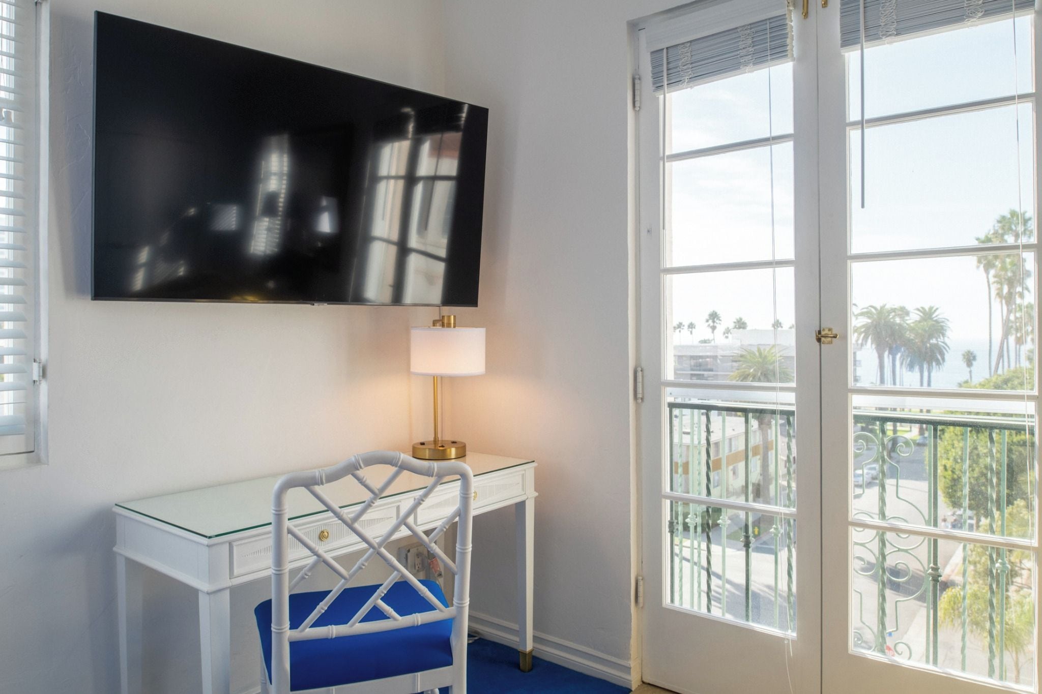 Una habitación con escritorio, una silla, televisor montado en la pared y un amplio ventanal con vistas a palmeras y edificios.