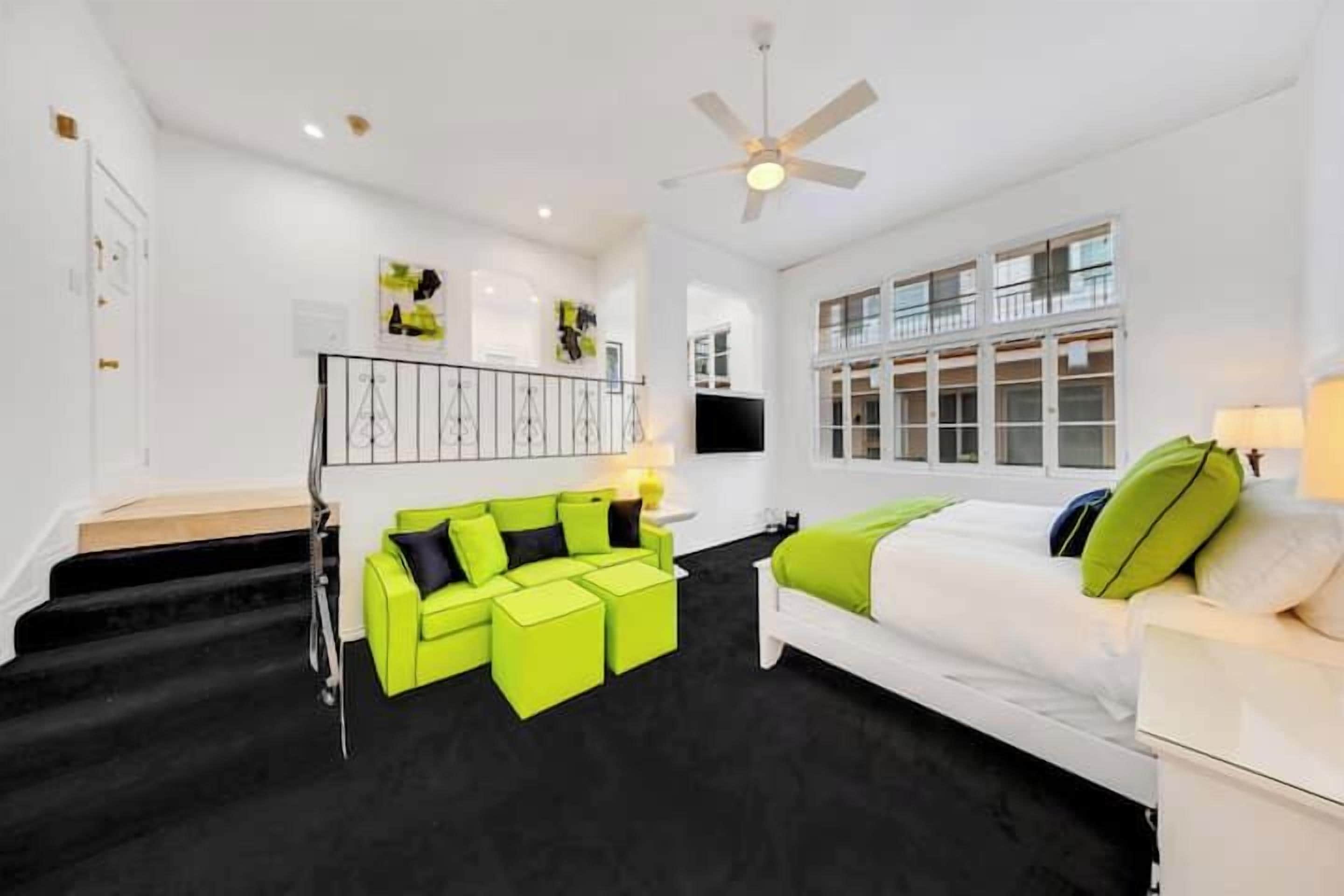 A modern hotel room with a black carpet, a white sofa with green cushions, a white bed, and a staircase leading to an upper level.