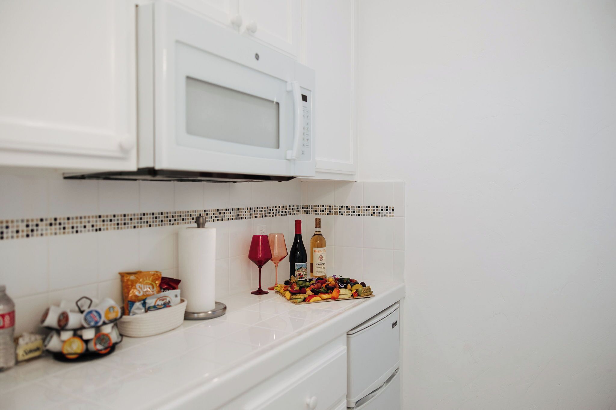A kitchen with white cabinets, a microwave, a tiled backsplash, and a countertop with snacks, wine glasses, and a bottle.