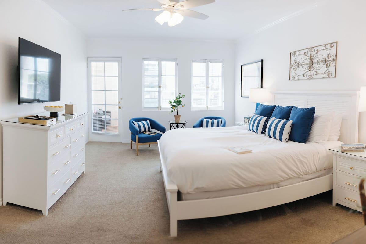 Un dormitorio con cama blanca, cómoda, televisor y ventilador de techo.