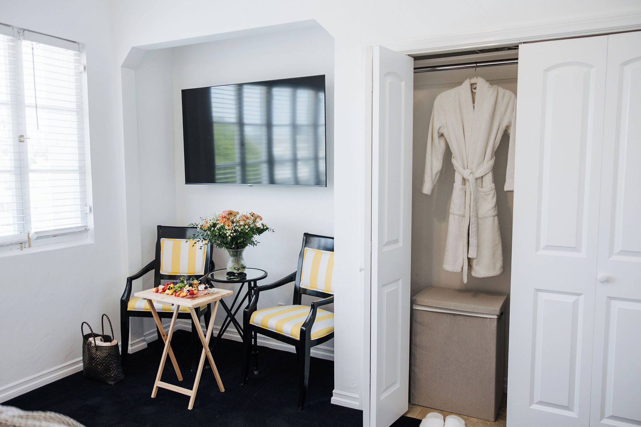 A room with a TV mounted on the wall, two chairs with striped cushions, a small table with a vase of flowers, and a closet with a robe hanging.