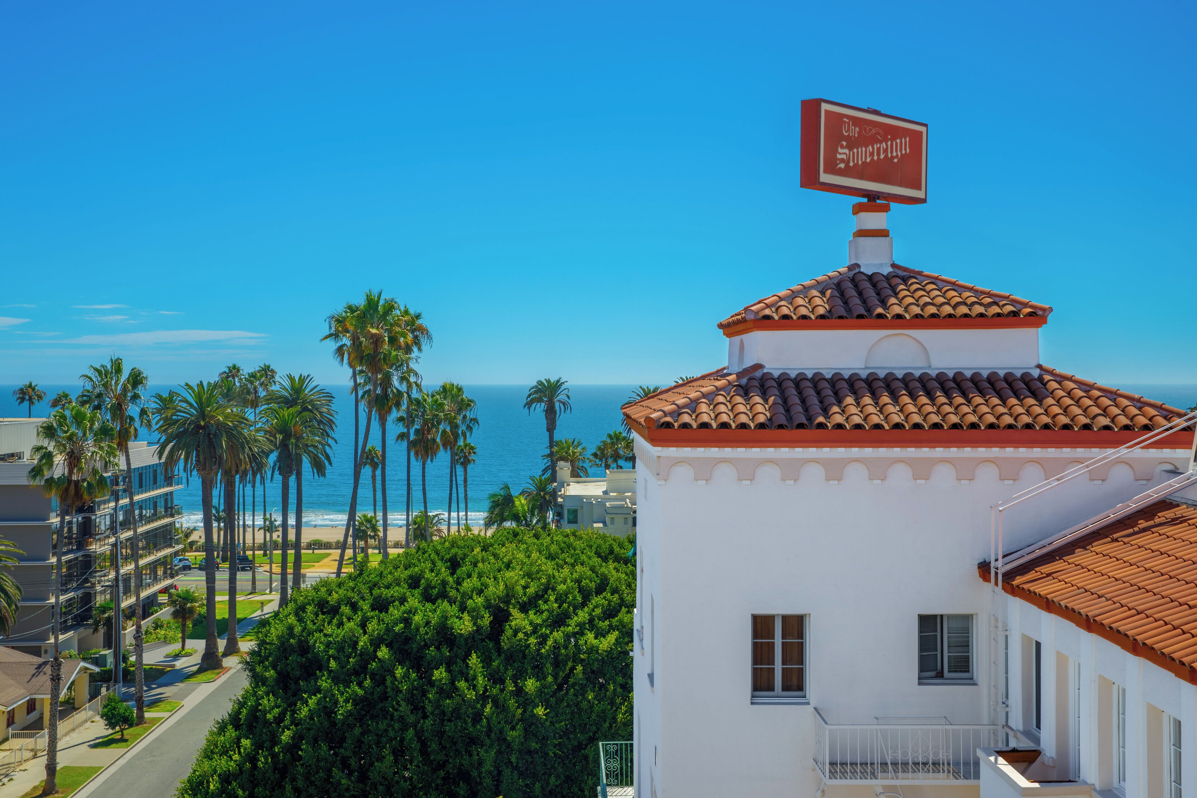 A white building with a red-tiled roof and a sign reading "The Soprano." It features a balcony and is surrounded by palm trees and a view of the ocean.