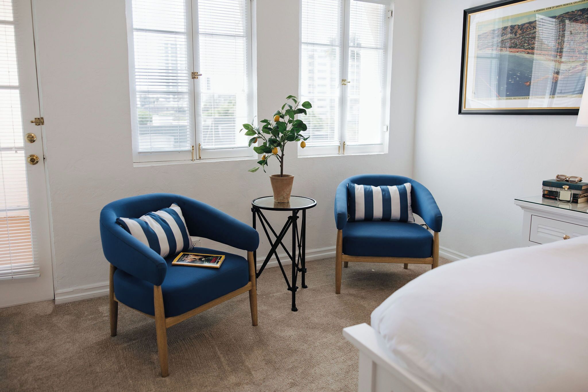 A room with two blue armchairs, a small table with a plant, a bed, and a framed picture on the wall.