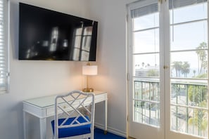 A room with a glass desk, a chair with a blue cushion, a mounted TV, and a door leading to a balcony with a view of palm trees.