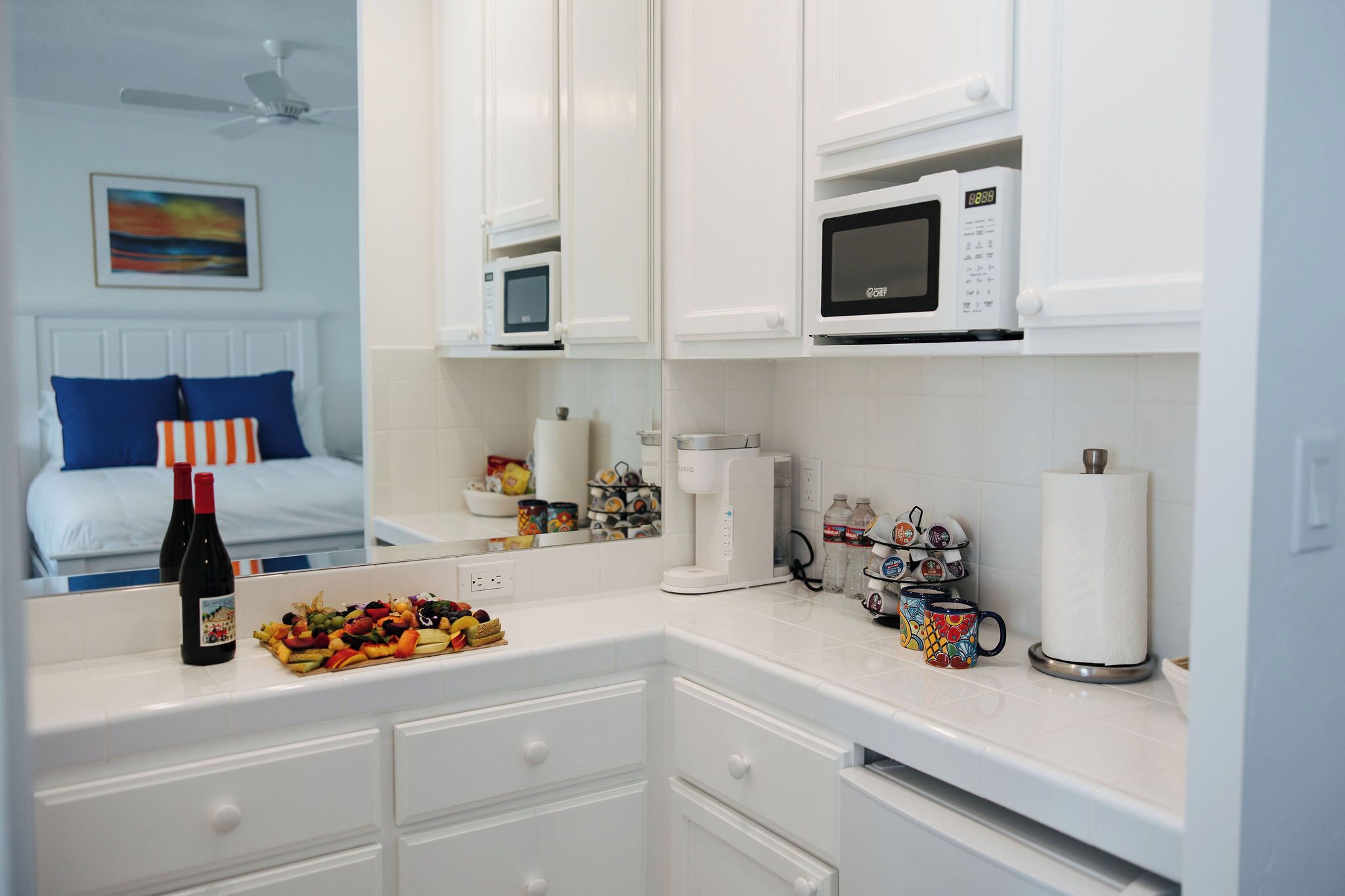 A modern kitchen with white cabinetry, a microwave, a coffee maker, and a breakfast bar with a fruit platter and wine bottles.