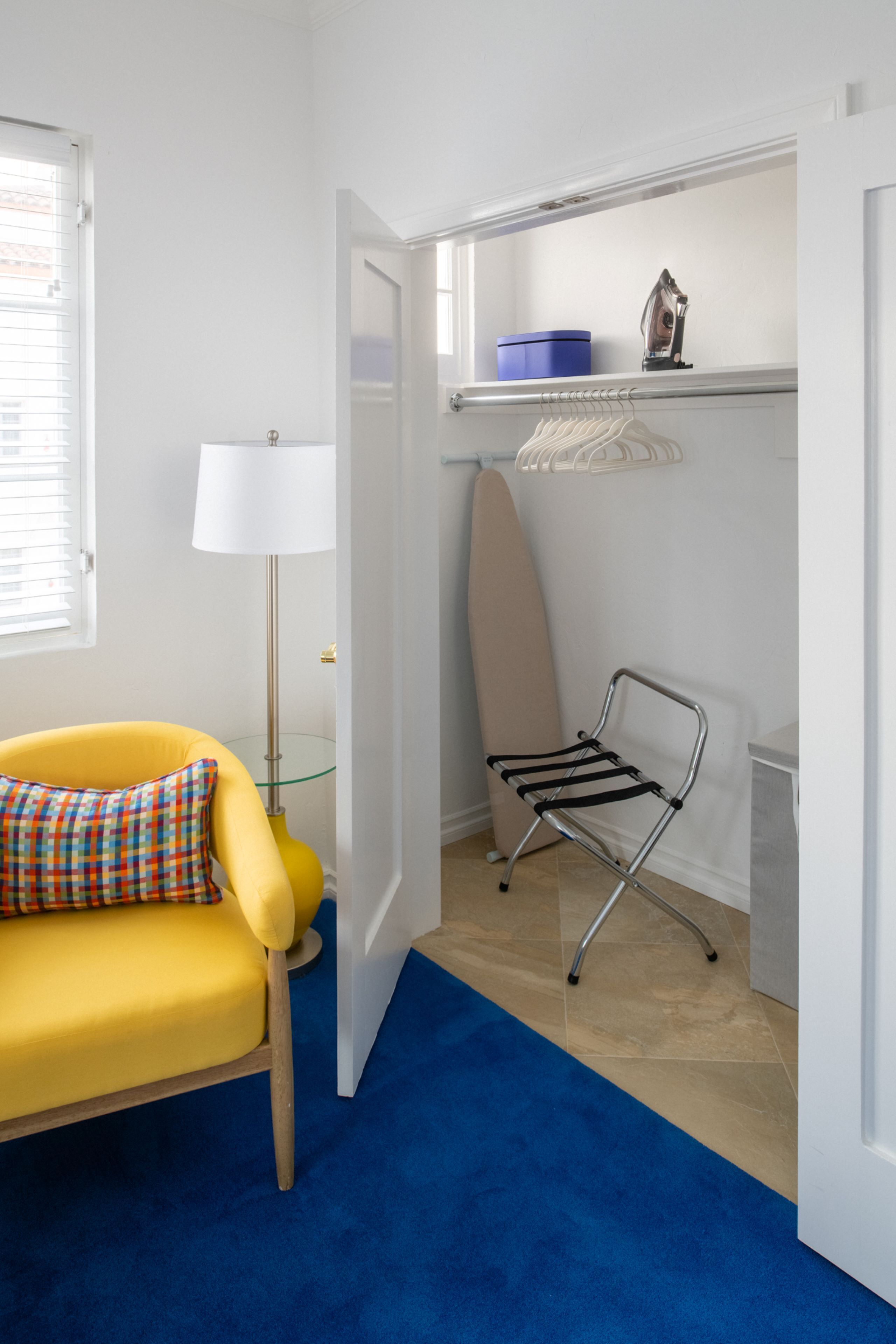 A compact room with a yellow armchair, a blue floor, a white lamp, a shelf with a blue container and an iron, and a hanging board.