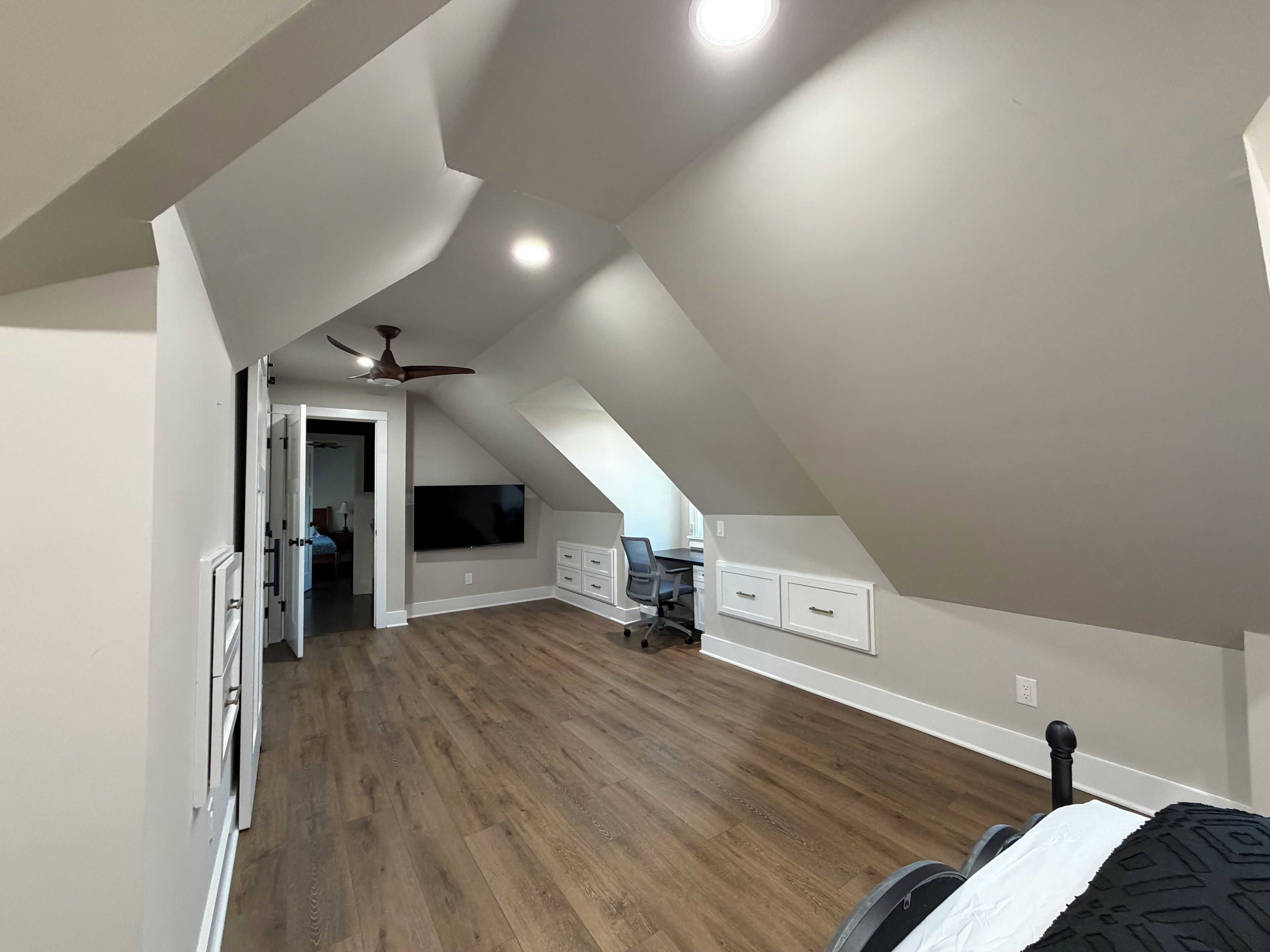 A spacious attic bedroom with a sloped ceiling, a ceiling fan, a flat-screen TV mounted on the wall, and a wooden floor.