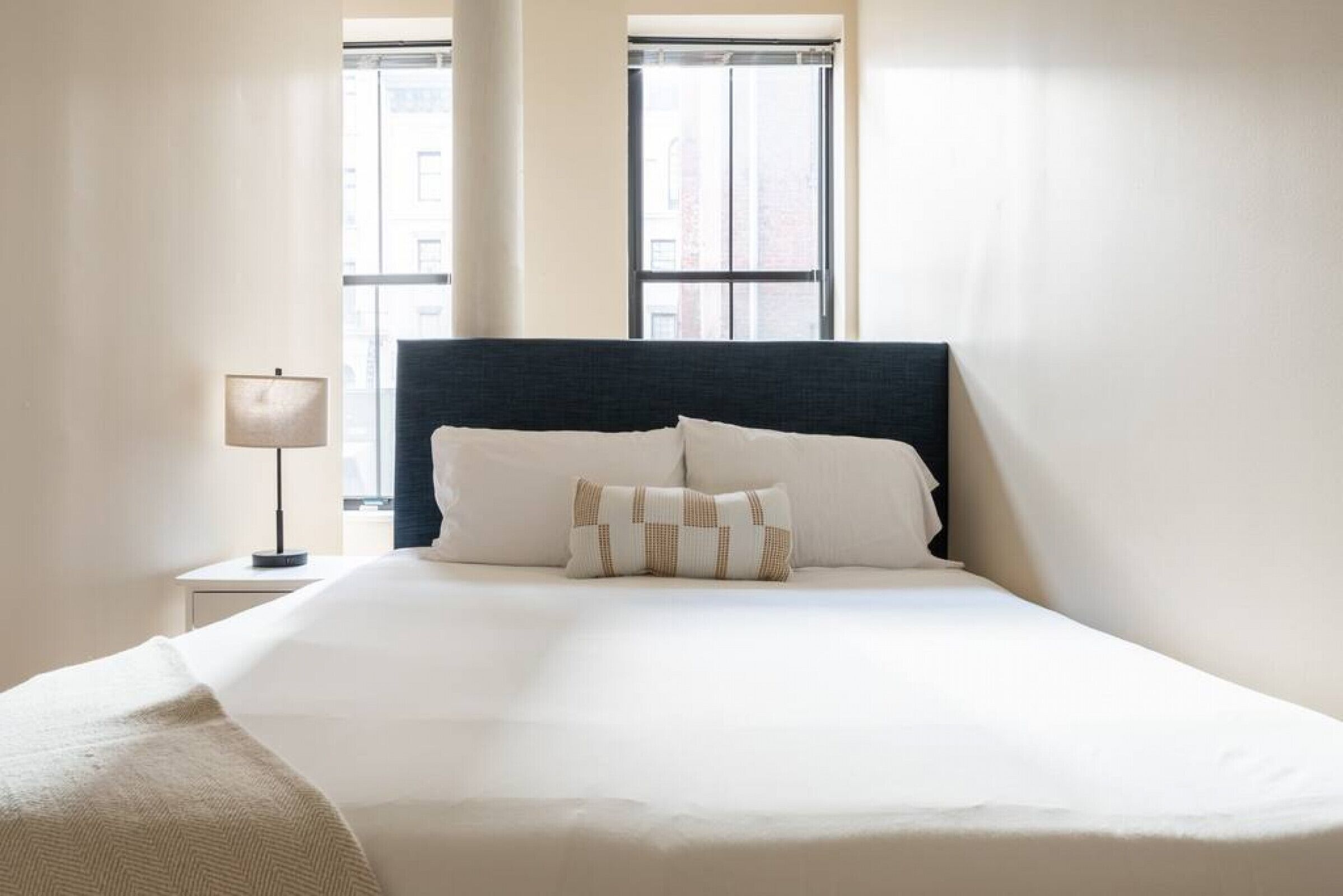 Een netjes opgemaakt bed met een donker hoofdbord, een bedlampje en een raam met jaloezieën.