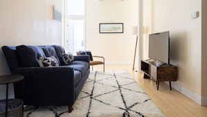 Un salon avec un canapé bleu foncé, un tapis blanc, un meuble TV en bois et un tableau encadré accroché au mur.