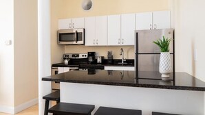 Une cuisine moderne avec des armoires blanches, un réfrigérateur en acier inoxydable, un four à micro-ondes et une machine à café. On y trouve trois tabourets noirs et une plante en pot sur le comptoir.