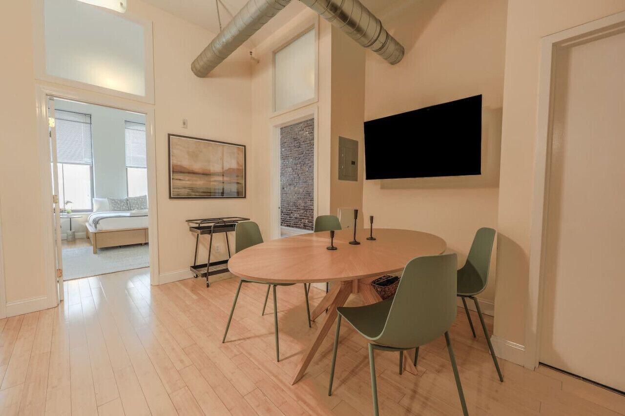 A modern living room with a round wooden table, green chairs, a flat-screen TV mounted on the wall, and a large abstract painting.
