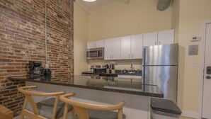 A compact kitchen with white cabinets, a stainless steel refrigerator, a microwave, and a coffee maker. There are three wooden chairs with cushions. - Stylish Exposed Brick 3BR! (Boston)