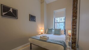 A bedroom with a bed, bedside lamps, a window, and a brick wall. - Stylish Exposed Brick 3BR! (Boston)