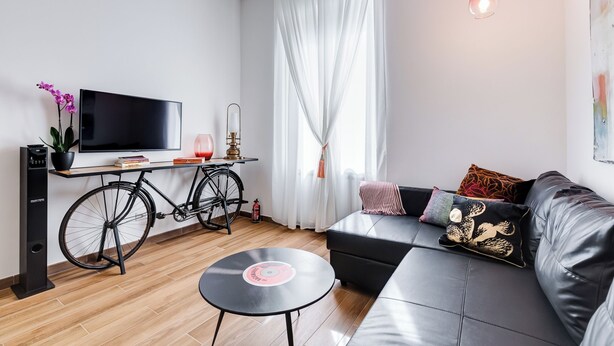 Una sala de estar moderna con un sofá negro, una mesita redonda, una bicicleta, un televisor y una ventana con cortinas translúcidas.