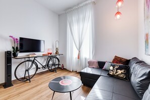 Egy modern nappali, melyben egy fekete kanapé, egy kis, kerek asztal, egy kerékpár, egy televízió és egy átlátszó, könnyű anyagból készült függönnyel ellátott ablak található.