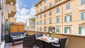 Un balcón con mobiliario de mimbre, una mesa con un croissant y vistas a un edificio histórico con contraventanas verdes.