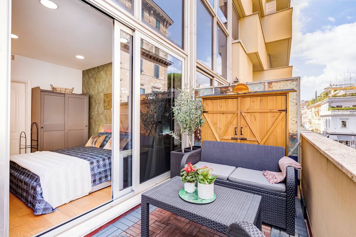 Un apartamento moderno con balcón, un sofá, una mesita con plantas en macetas y un ventanal corredero que da a un dormitorio.