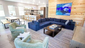 A modern living room with a sectional sofa, a coffee table, and two accent chairs. A kitchen is visible in the background with a wooden accent wall and a painting. - Firefly South: 3,800 sqft - Sleeps 16! 2-min walk to pool and beach! (Saint Teresa)