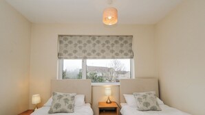 A bedroom with two beds, a window with a view, a bedside table with a lamp, and a ceiling light. - Lavender Harmony House (Swindon)