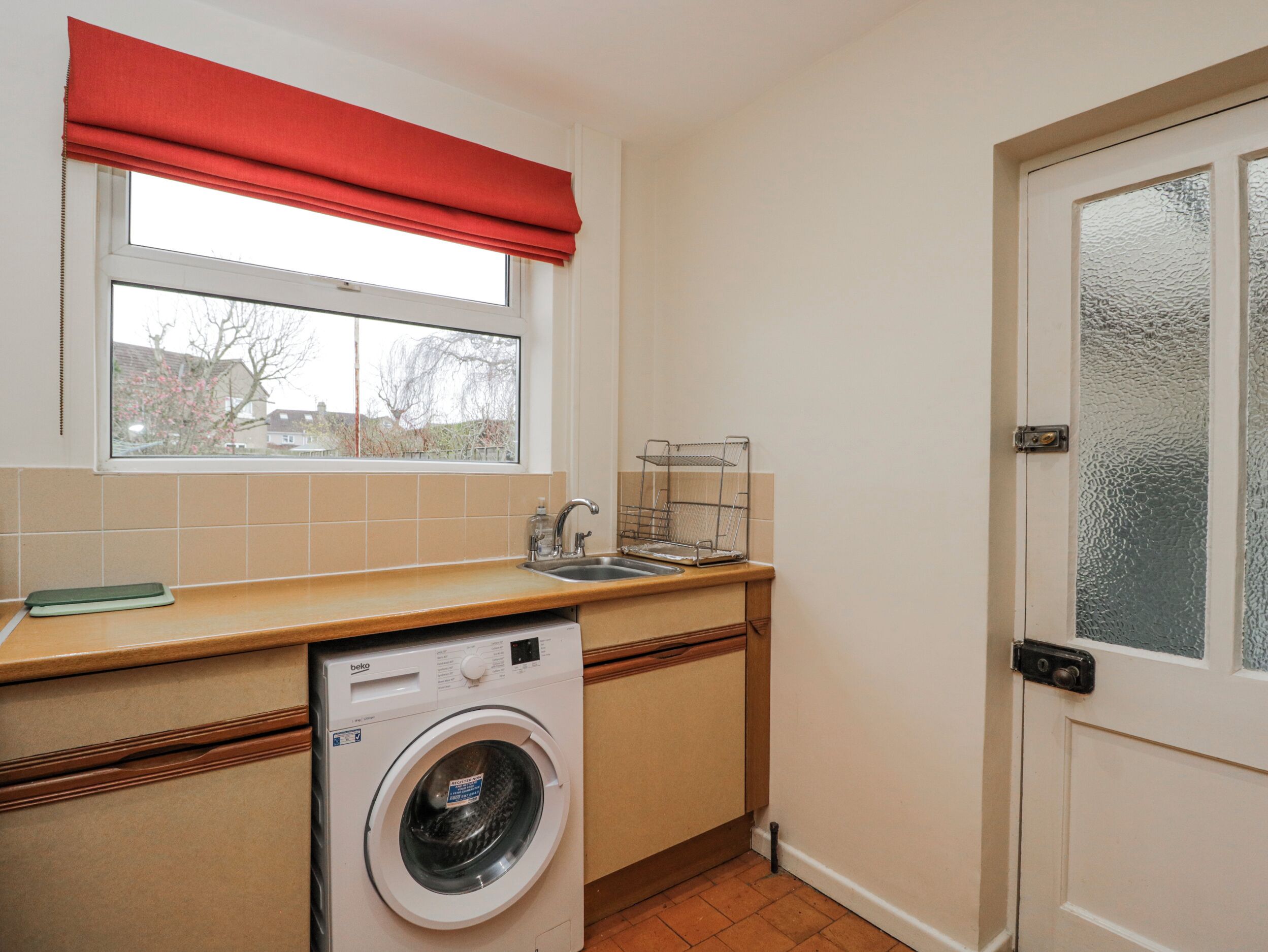 Een kleine keuken met een wasmachine, een raam met een rode jaloezie en een deur met geëtste glas.