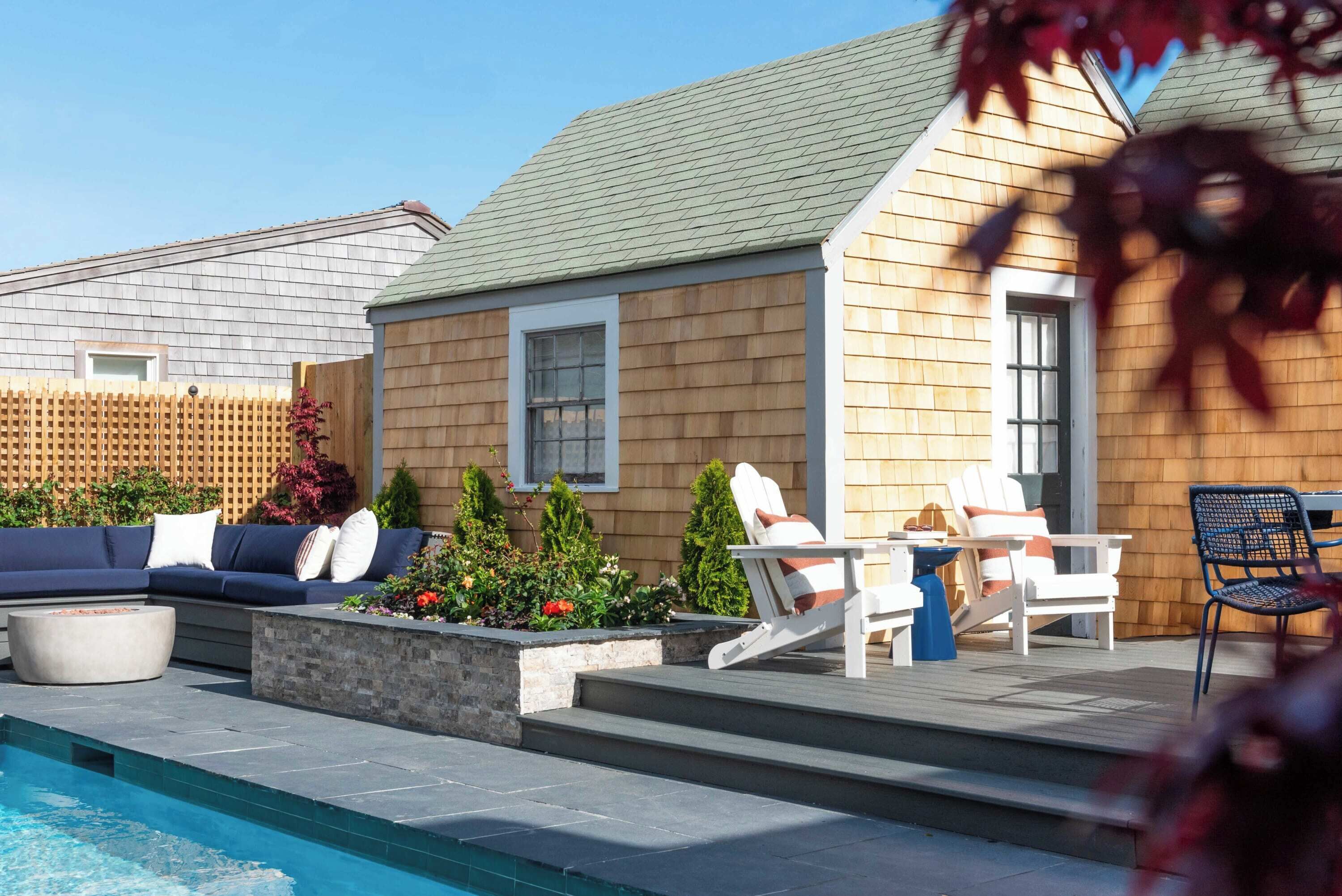 A poolside area with a wooden deck, a seating area with a sofa and chairs, and a small building with a window.