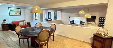 A dining area with a round table and chairs, a red sofa, and a kitchen area with a window and a shelf with cups.