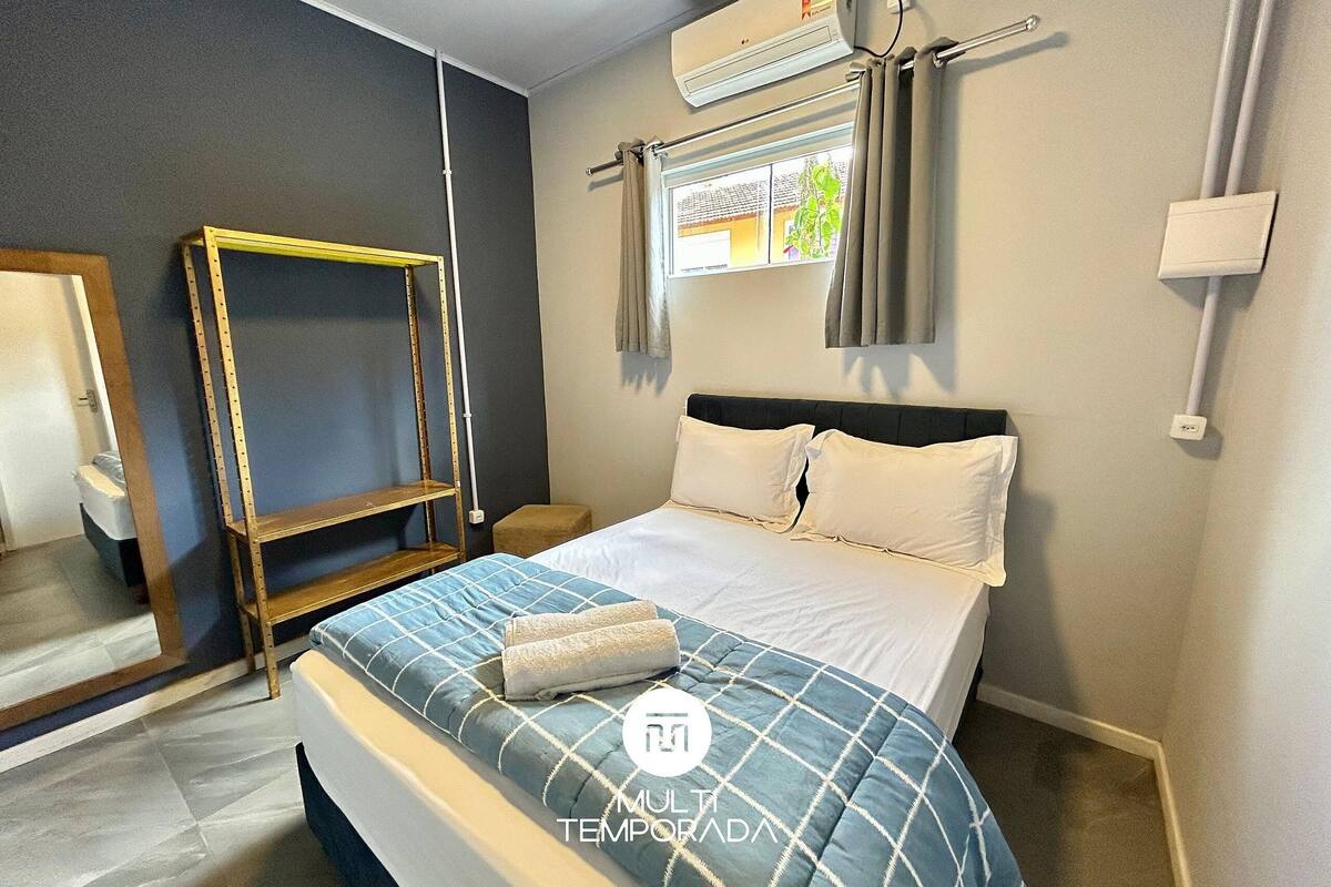 A bedroom with a bed, a window with curtains, an air conditioning unit, and a wooden shelf.
