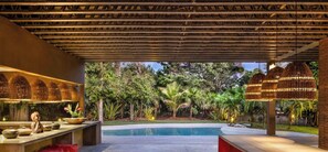 Un espace piscine extérieur doté d’un coin salon couvert, entouré d’une végétation luxuriante et surmonté d’une structure en bois.