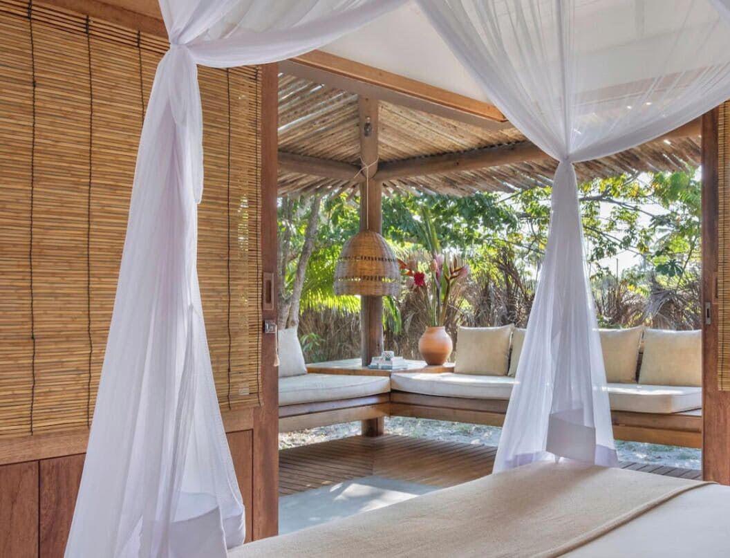 Una habitación con cama, zona de estar con cojines, lámpara colgante y vistas al exterior con vegetación.