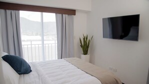 A hotel room with a bed, a TV mounted on the wall, a window with curtains, and a potted plant. - Glam | Modern apartment in exclusive area of Xela (Quetzaltenango)