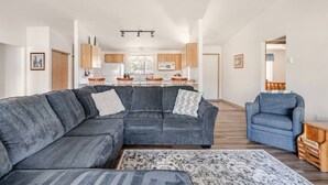 A modern living room with a sectional sofa, a blue armchair, and a patterned rug. A kitchen is visible in the background with wooden cabinets and a dining area. - Adventure Retreat - Sunriver/Bachelor/Waterfront (Bend)