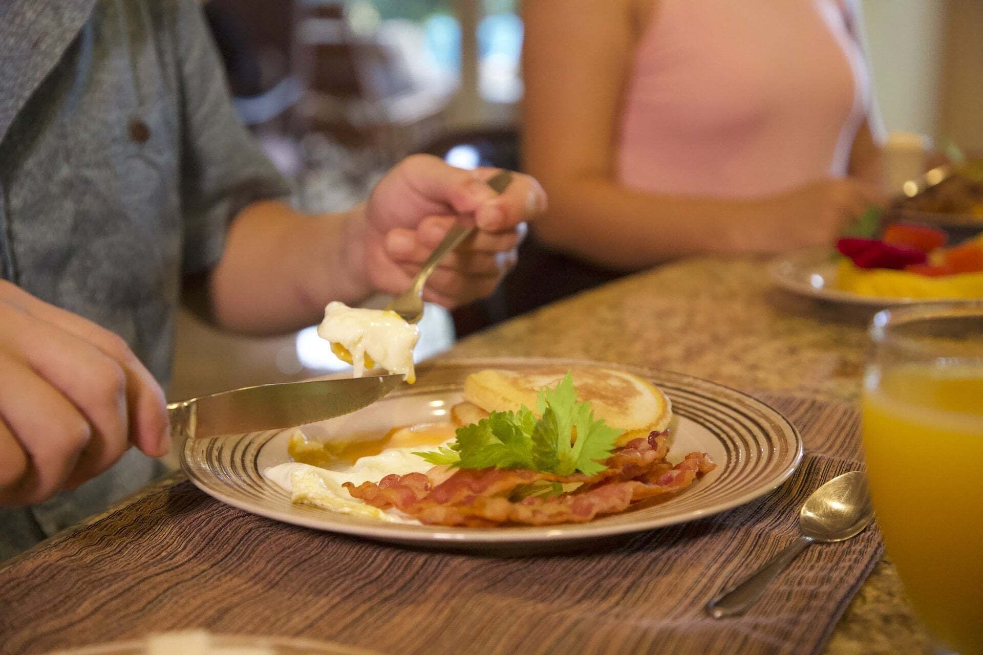 A breakfast setting with a plate of eggs, bacon, and a garnish, a spoon, and a glass of orange juice on a wooden table.